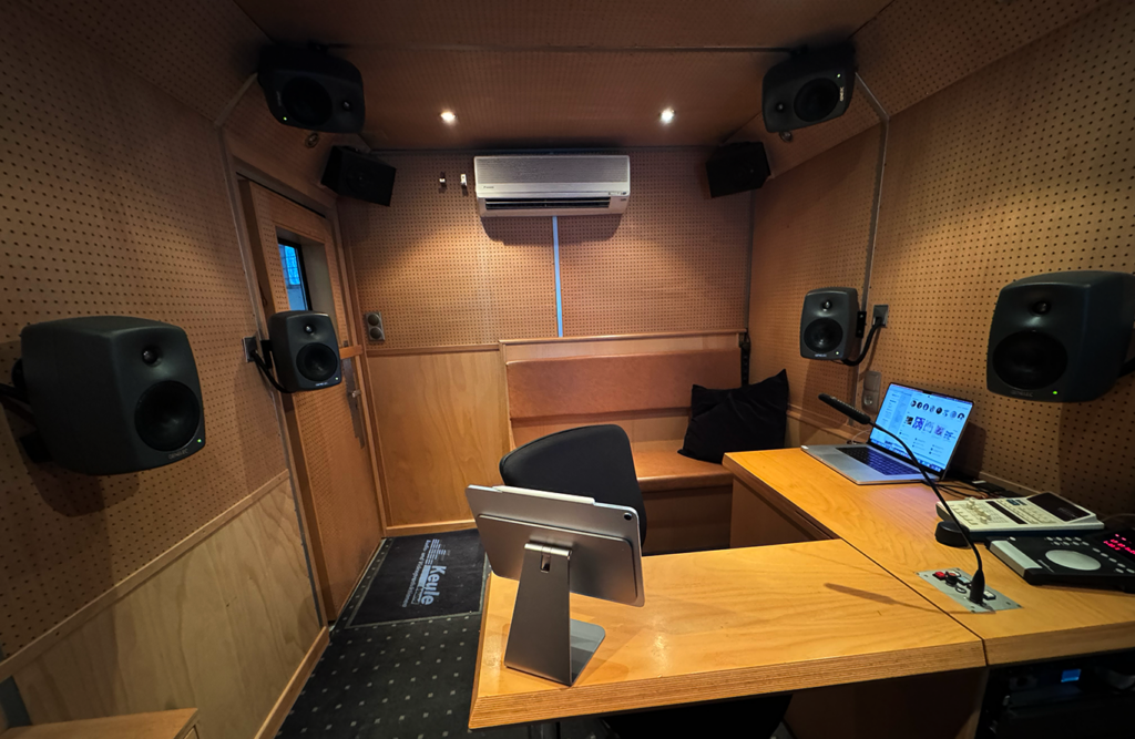 Im Vordergrund ein Holztisch mit einem Display Rechts davon zwei Bedienungseinheiten von Hallgeräten Rundherum um den Platz Lautsprecher der Marke Genelec für Dolby Atmos. Zentral der Bürostuhl. Im Hintergrund eine Sitzbank mit Leder bezogen für Artist oder Producer.